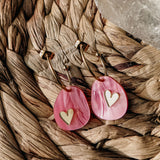 Gold lightweight huggie hoops with pink marbled acrylic, teardrop-shaped pendants featuring gold heart inlays, rest on a woven natural fiber surface beside a translucent crystal—a romantic touch from the Valentine Pebble Earrings collection.