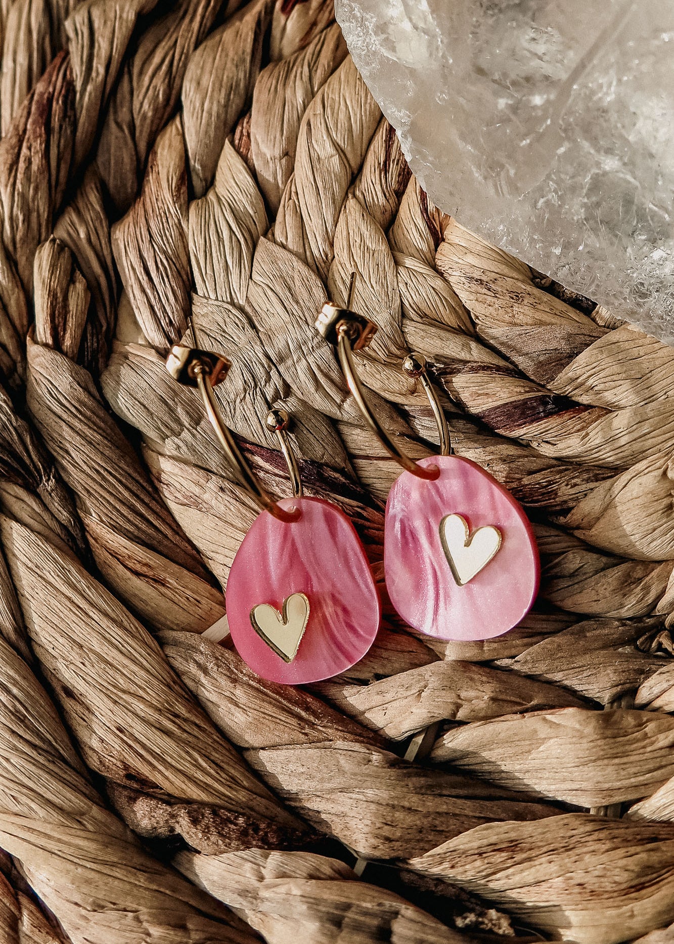 Gold lightweight huggie hoops with pink marbled acrylic, teardrop-shaped pendants featuring gold heart inlays, rest on a woven natural fiber surface beside a translucent crystal—a romantic touch from the Valentine Pebble Earrings collection.