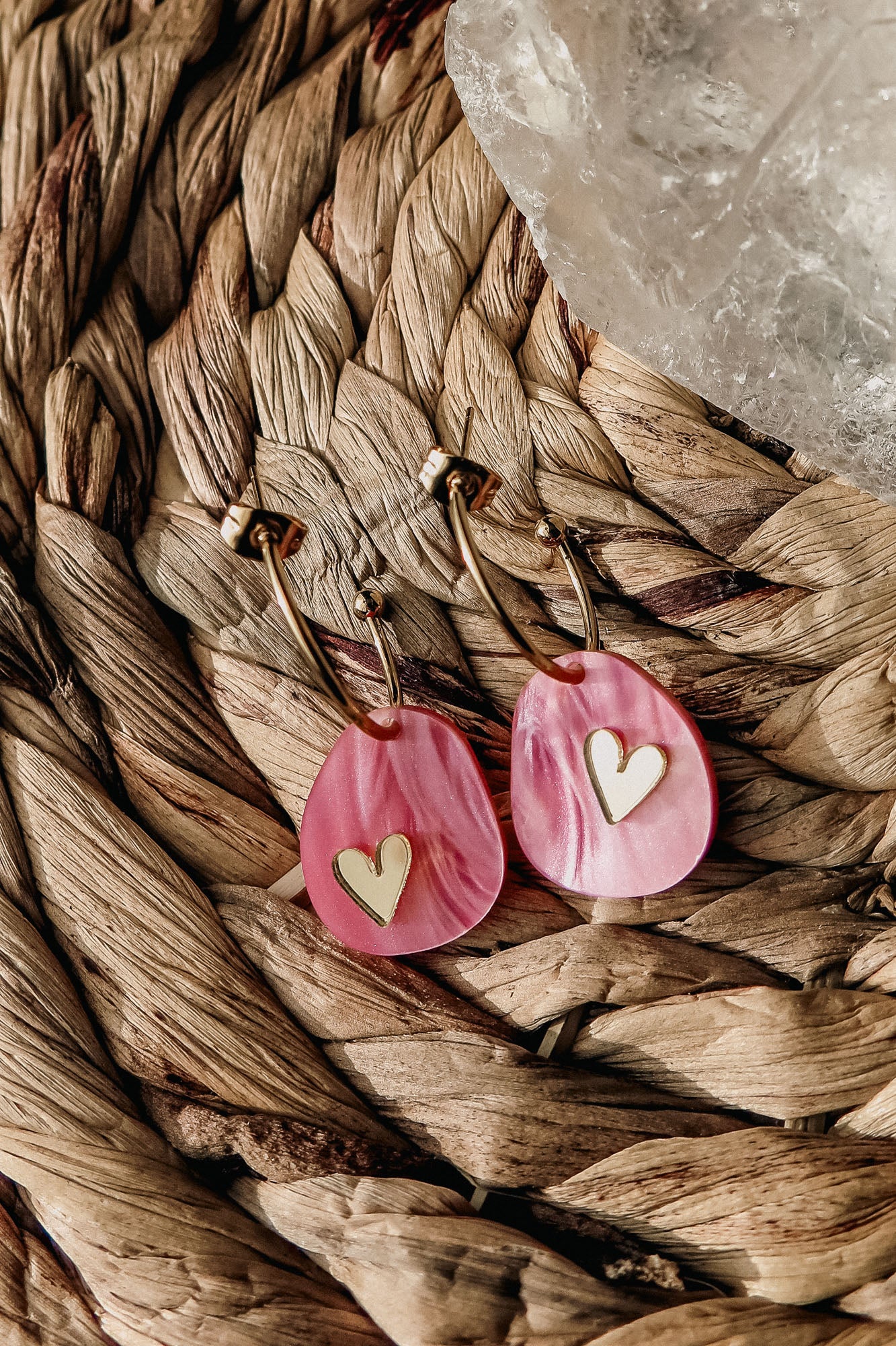 Gold lightweight huggie hoops with pink marbled acrylic, teardrop-shaped pendants featuring gold heart inlays, rest on a woven natural fiber surface beside a translucent crystal—a romantic touch from the Valentine Pebble Earrings collection.