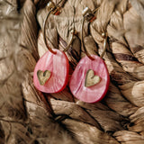 A pair of lightweight huggie hoops featuring pink marbled acrylic oval pendants and small gold heart accents, displayed on a woven, textured surface—meet the charming Valentine Pebble Earrings.