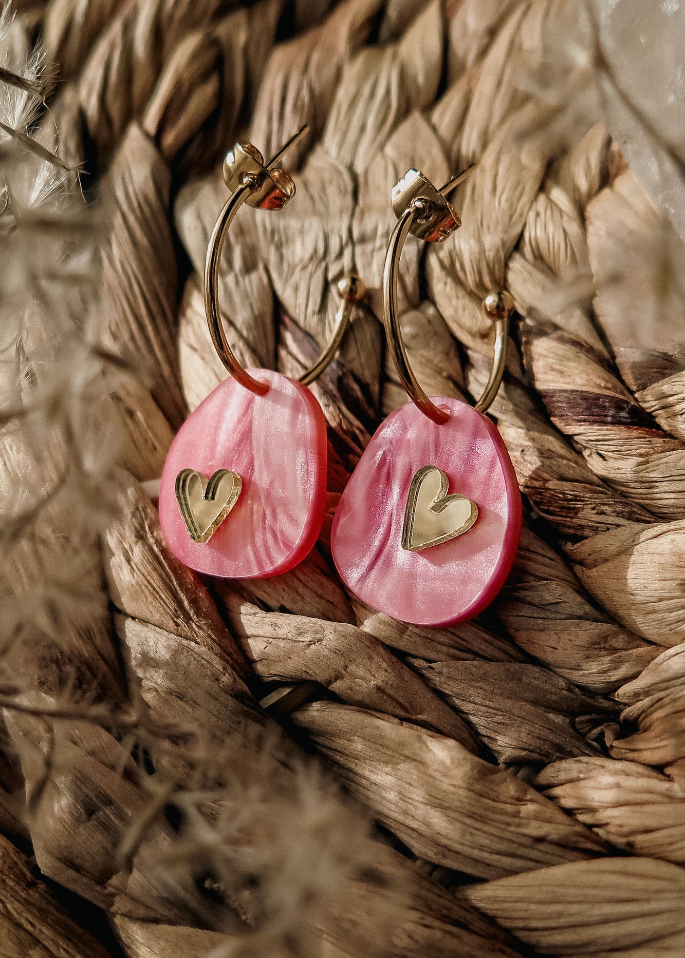 A pair of lightweight huggie hoops featuring pink marbled acrylic oval pendants and small gold heart accents, displayed on a woven, textured surface—meet the charming Valentine Pebble Earrings.