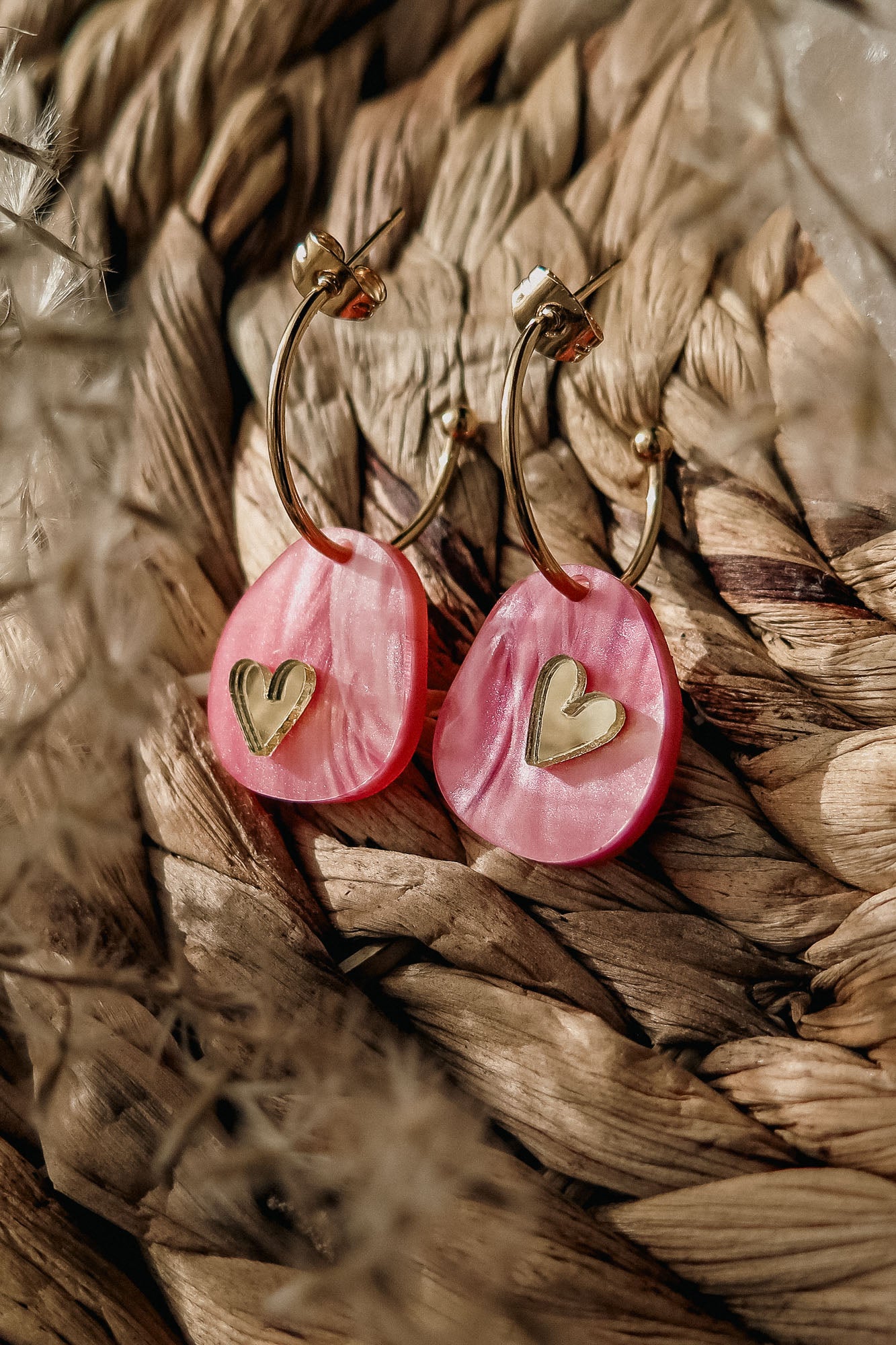 A pair of lightweight huggie hoops featuring pink marbled acrylic oval pendants and small gold heart accents, displayed on a woven, textured surface—meet the charming Valentine Pebble Earrings.