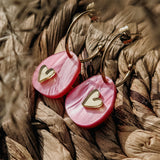 A close-up of lightweight huggie hoops with pink marbled acrylic pendants, each featuring a small gold heart, resting on a woven natural fiber surface—perfect for fans of the Valentine Pebble Earrings.