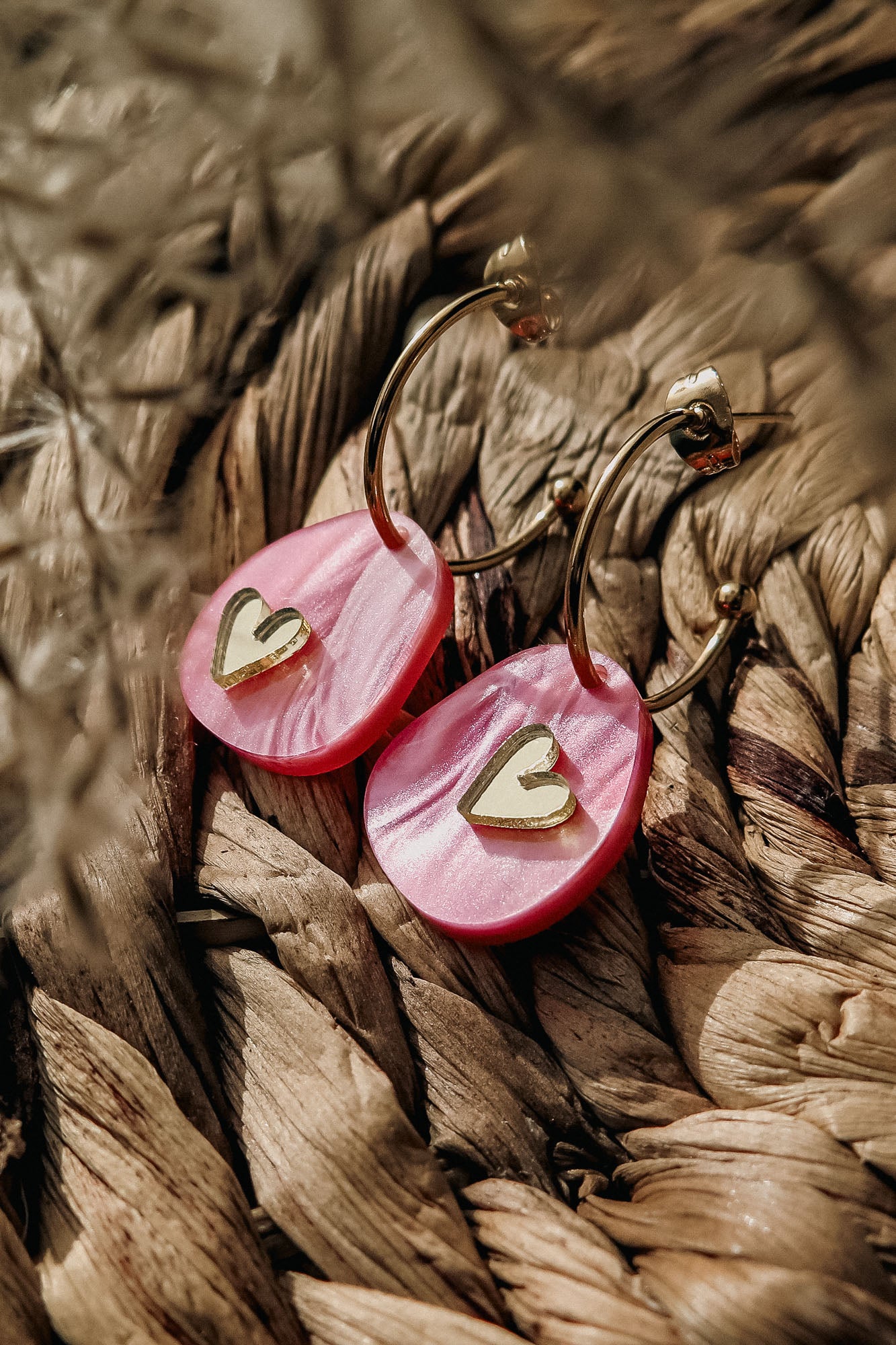 A close-up of lightweight huggie hoops with pink marbled acrylic pendants, each featuring a small gold heart, resting on a woven natural fiber surface—perfect for fans of the Valentine Pebble Earrings.