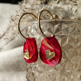 Gold hoop earrings with red and pink marbled resin charms, each adorned with a small gold heart, create a romantic accessory. Displayed on a textured glass container, these are the perfect Valentine Pebble Earrings for a love-inspired style.