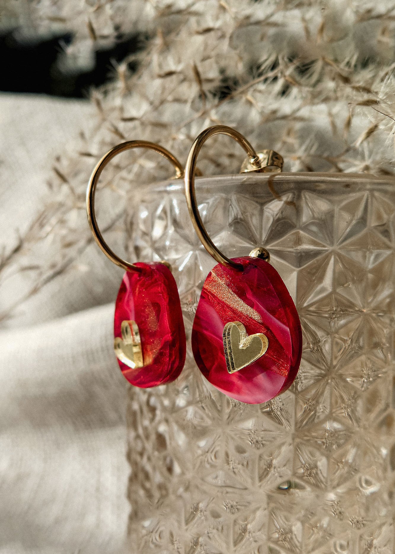 Gold hoop earrings with red and pink marbled resin charms, each adorned with a small gold heart, create a romantic accessory. Displayed on a textured glass container, these are the perfect Valentine Pebble Earrings for a love-inspired style.