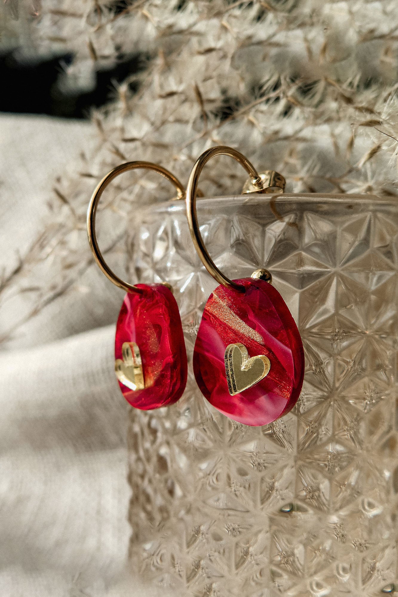 Gold hoop earrings with red and pink marbled resin charms, each adorned with a small gold heart, create a romantic accessory. Displayed on a textured glass container, these are the perfect Valentine Pebble Earrings for a love-inspired style.