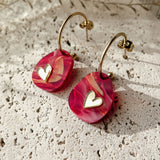 A pair of gold hoop earrings with pink, marbled resin drops and small white heart charms, the Valentine Pebble Earrings are a romantic accessory displayed on a textured stone surface under natural light.