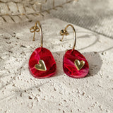 A pair of gold hoop earrings with red, irregular-shaped pendants featuring small gold heart accents, the Valentine Pebble Earrings are a romantic accessory displayed on a textured light stone surface with dried plants in the background.