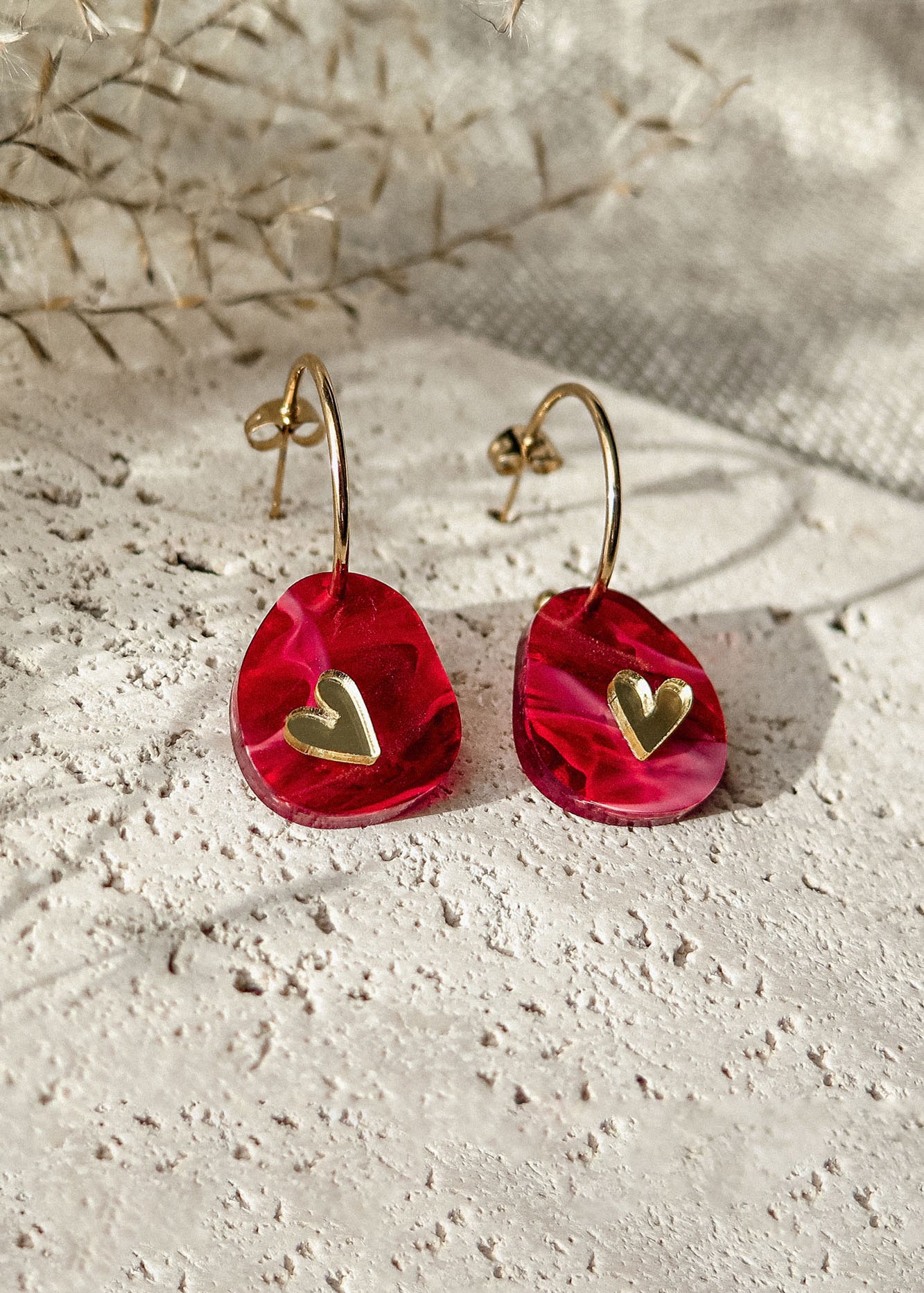 A pair of gold hoop earrings with red, irregular-shaped pendants featuring small gold heart accents, the Valentine Pebble Earrings are a romantic accessory displayed on a textured light stone surface with dried plants in the background.