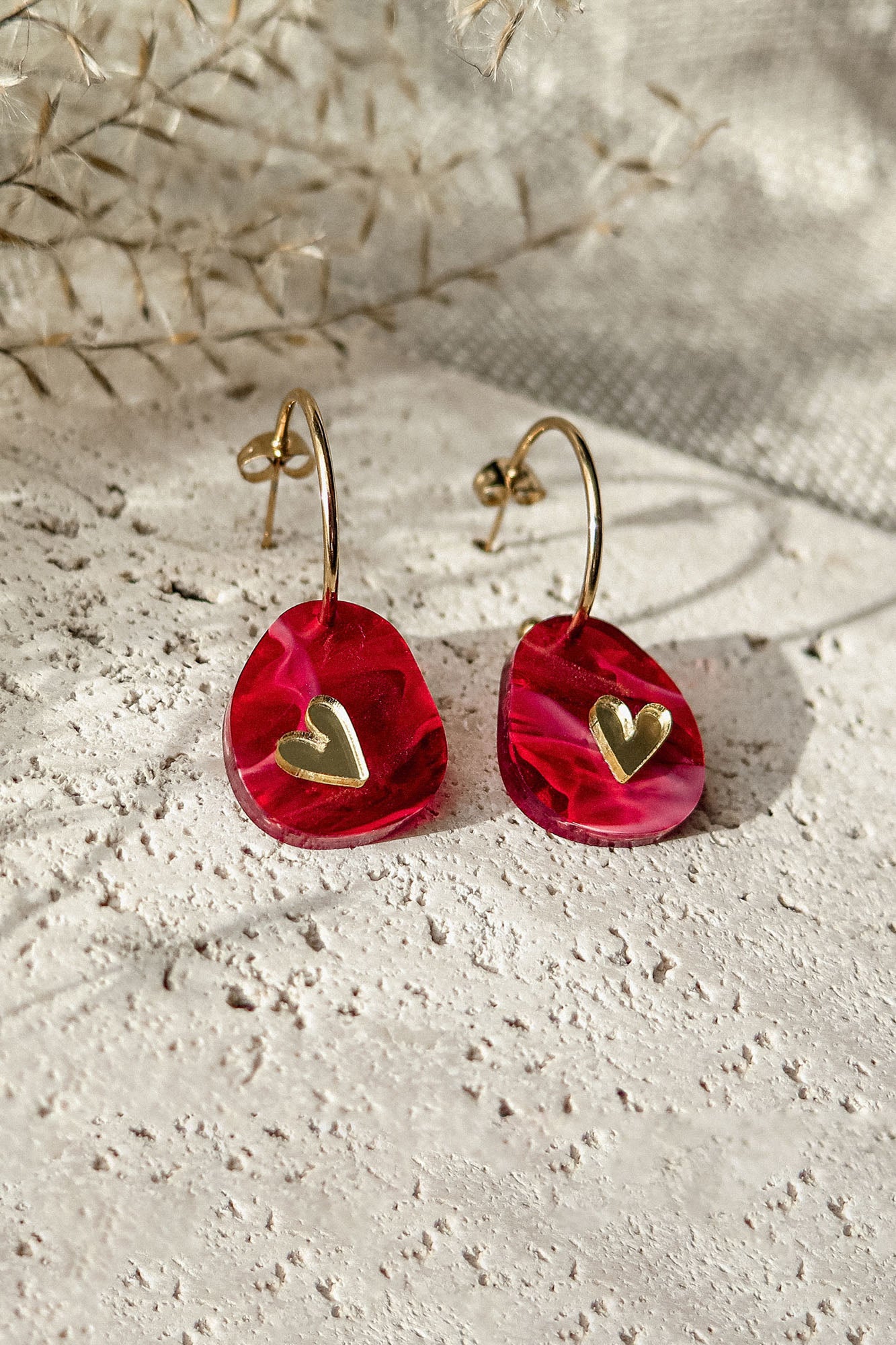 A pair of gold hoop earrings with red, irregular-shaped pendants featuring small gold heart accents, the Valentine Pebble Earrings are a romantic accessory displayed on a textured light stone surface with dried plants in the background.