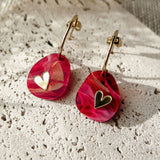 A pair of gold hoop Valentine Pebble Earrings with marbled pink resin pendants, each featuring a small gold heart detail, resting on a textured white stone surface—a romantic accessory perfect for love-inspired style.