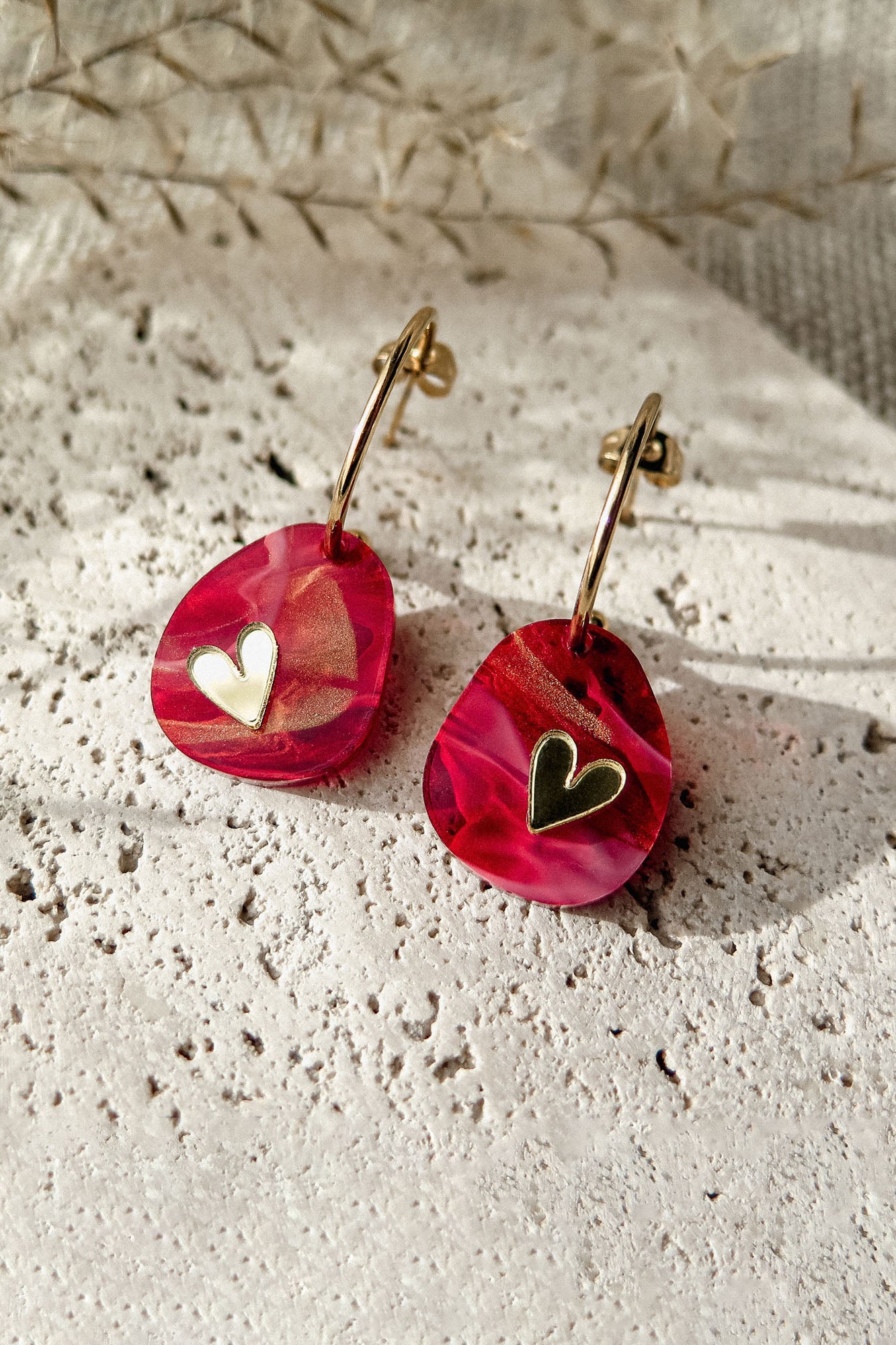 A pair of gold hoop Valentine Pebble Earrings with marbled pink resin pendants, each featuring a small gold heart detail, resting on a textured white stone surface—a romantic accessory perfect for love-inspired style.
