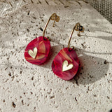 A pair of Valentine Pebble Earrings: gold hoops with red and pink marbled teardrop pendants and small gold heart charms, displayed on textured stone with dried grass—a perfect romantic accessory for a love-inspired style.