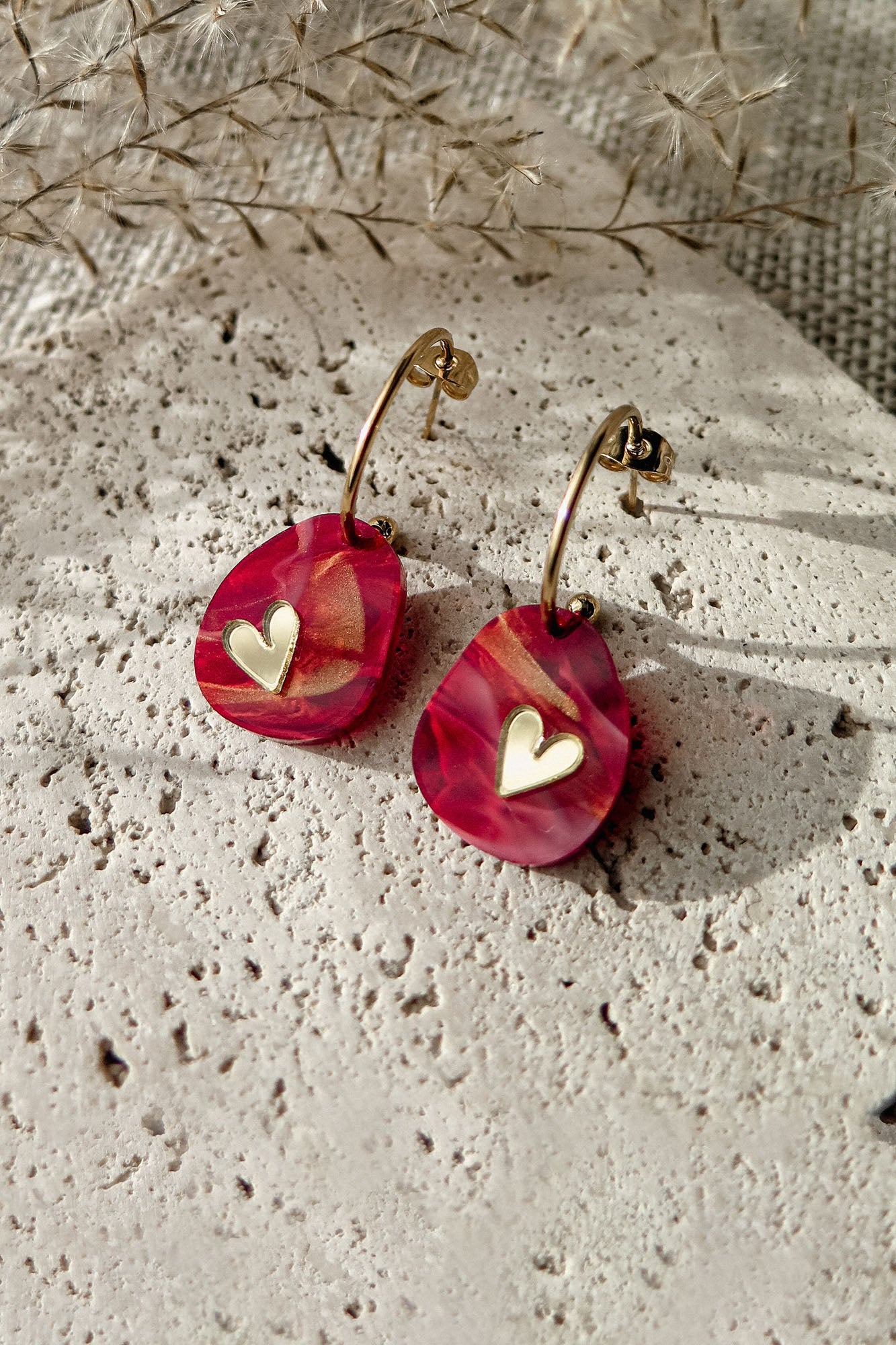 A pair of Valentine Pebble Earrings: gold hoops with red and pink marbled teardrop pendants and small gold heart charms, displayed on textured stone with dried grass—a perfect romantic accessory for a love-inspired style.