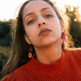 A woman with long brown hair wearing a red sweater and mismatched earrings in bold colors looks confidently at the camera outdoors, with sunlight and greenery in the background.