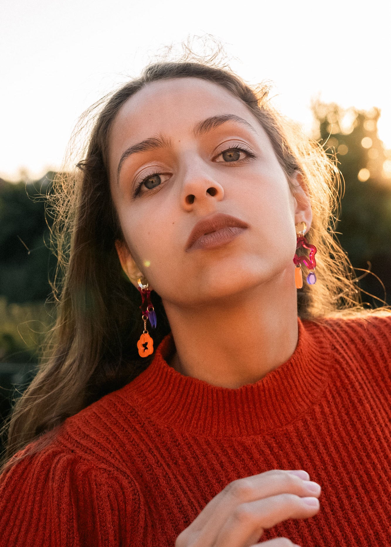 A woman with long brown hair wearing a red sweater and mismatched earrings in bold colors looks confidently at the camera outdoors, with sunlight and greenery in the background.
