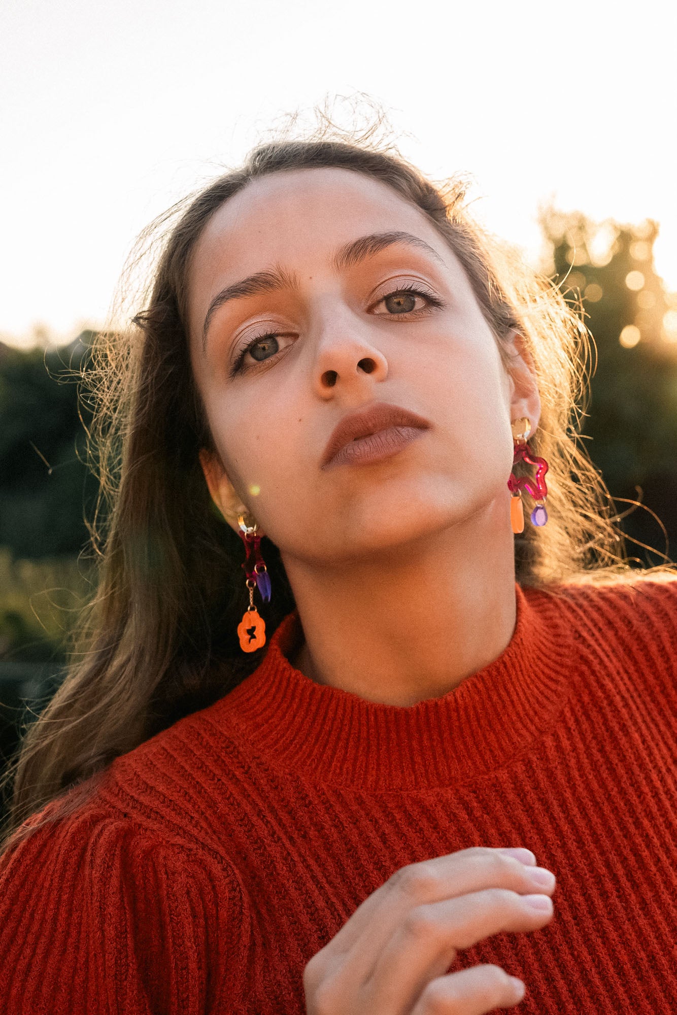 A woman with long brown hair wearing a red sweater and mismatched earrings in bold colors looks confidently at the camera outdoors, with sunlight and greenery in the background.