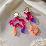 Two colorful, abstract earrings with bold colors rest on soft white fabric. Each earring features gold hardware and abstract shapes in wavy pink acrylic, translucent purple, and coral orange—making this a striking pair of mismatched earrings. Dried white plants appear nearby.
