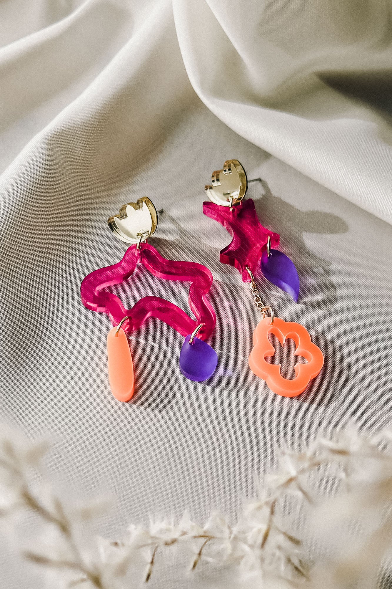 Two colorful, abstract earrings with bold colors rest on soft white fabric. Each earring features gold hardware and abstract shapes in wavy pink acrylic, translucent purple, and coral orange—making this a striking pair of mismatched earrings. Dried white plants appear nearby.