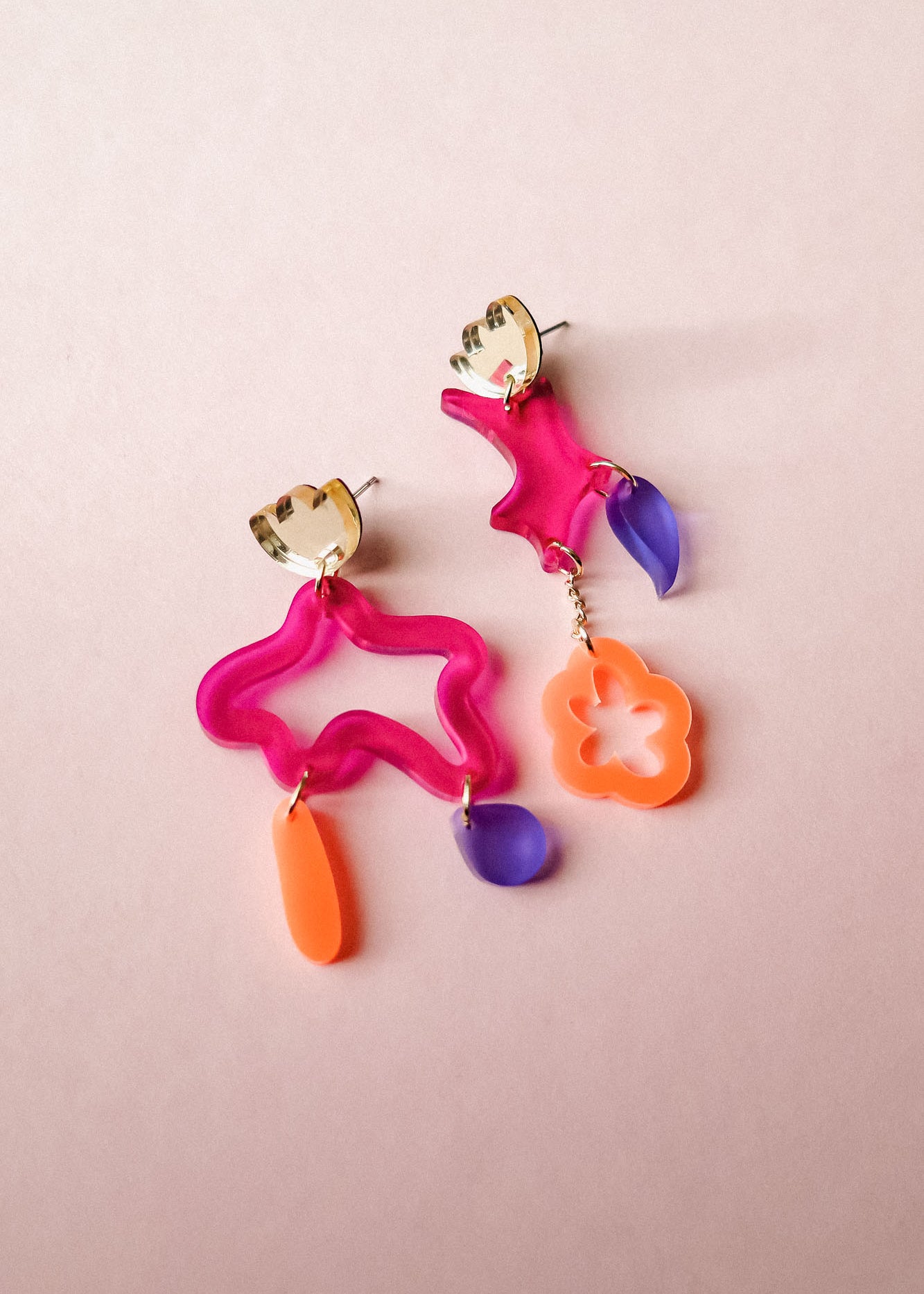 Two mismatched earrings in bold colors of pink, orange, and purple acrylic, featuring playful abstract shapes. Each unique earring dangles with geometric forms, making these Mismatched Earrings a vibrant statement on a light pink background.