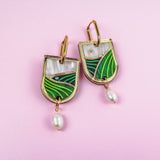 A pair of Vineyard Inspired Art Nouveau Dangle Earrings with green and white enamel landscape designs, accented with pearls & rhinestones and hanging pearl drops, displayed on a light pink background.
