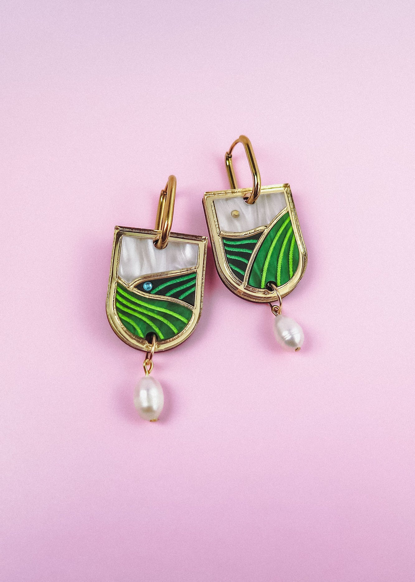 A pair of Vineyard Inspired Art Nouveau Dangle Earrings with green and white enamel landscape designs, accented with pearls & rhinestones and hanging pearl drops, displayed on a light pink background.