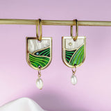 Gold Vineyard Inspired Art Nouveau Dangle Earrings with green and white enamel, an abstract landscape design, and small pearl drops, hang from a horizontal rod against a soft pink and white background.