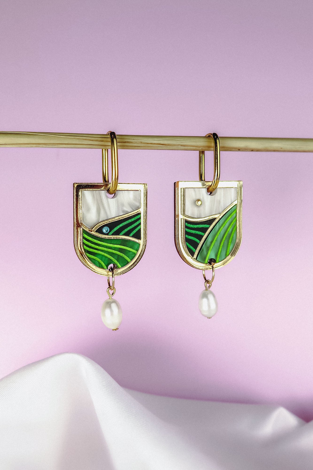 Gold Vineyard Inspired Art Nouveau Dangle Earrings with green and white enamel, an abstract landscape design, and small pearl drops, hang from a horizontal rod against a soft pink and white background.