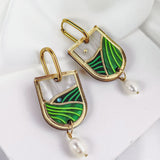 A pair of Vineyard Inspired Art Nouveau Dangle Earrings featuring abstract green enamel designs, pearls & rhinestones, and a dangling white pearl, beautifully displayed on a white fabric background.
