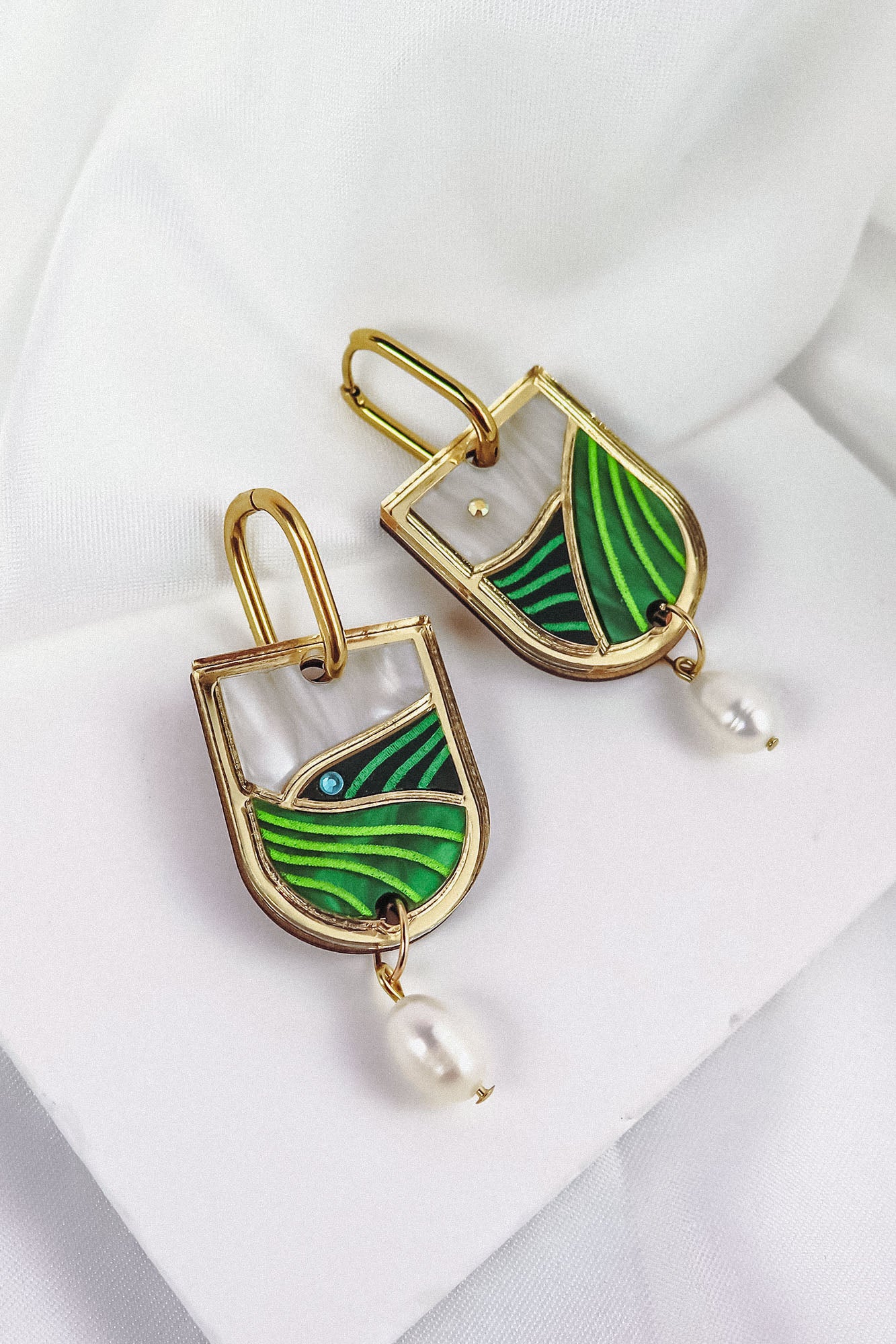A pair of Vineyard Inspired Art Nouveau Dangle Earrings featuring abstract green enamel designs, pearls & rhinestones, and a dangling white pearl, beautifully displayed on a white fabric background.