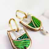 Close-up of Vineyard Inspired Art Nouveau Dangle Earrings, featuring green and white enamel designs, a touch of blue, and a dangling white pearl, displayed on a white surface.