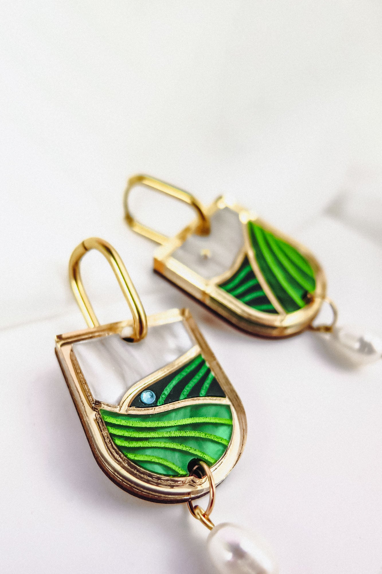 Close-up of Vineyard Inspired Art Nouveau Dangle Earrings, featuring green and white enamel designs, a touch of blue, and a dangling white pearl, displayed on a white surface.
