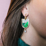 A close-up of a person wearing Vineyard Inspired Art Nouveau Dangle Earrings, featuring green, white, and silver enamel details with pearls & rhinestones. The person has brown hair and is wearing a dark green top.