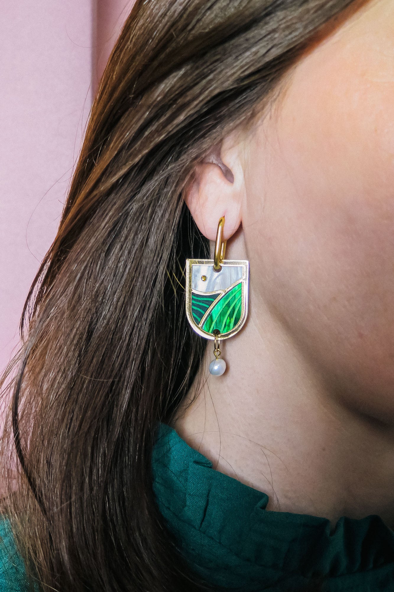 A close-up of a person wearing Vineyard Inspired Art Nouveau Dangle Earrings, featuring green, white, and silver enamel details with pearls & rhinestones. The person has brown hair and is wearing a dark green top.