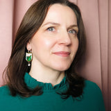 A woman with shoulder-length brown hair, wearing a teal top with a ruffled collar and elegant Vineyard Inspired Art Nouveau Dangle Earrings adorned with pearls & rhinestones, stands in front of pink curtains, looking slightly to the side and smiling softly.