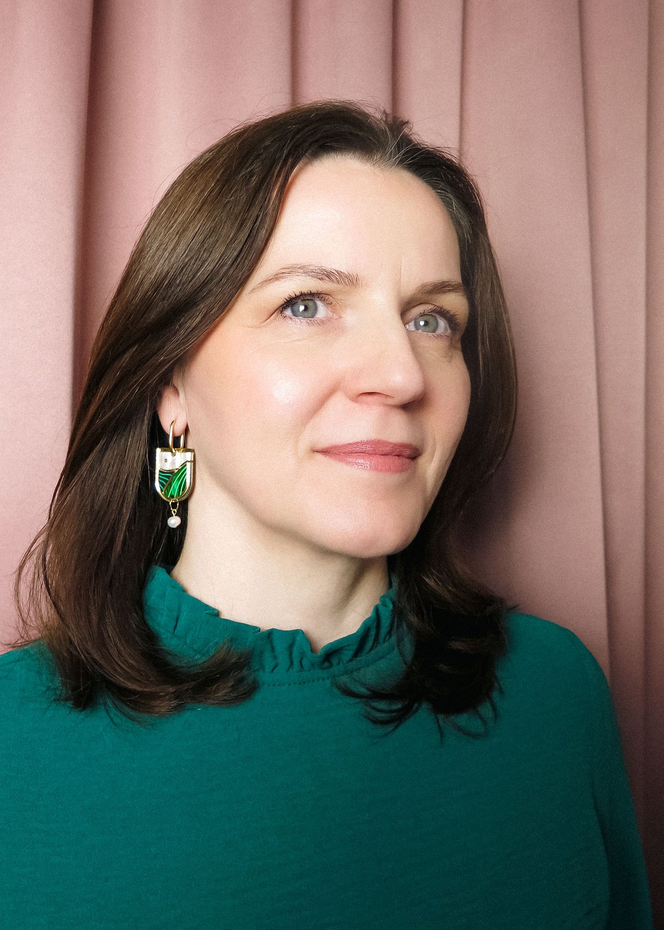 A woman with shoulder-length brown hair, wearing a teal top with a ruffled collar and elegant Vineyard Inspired Art Nouveau Dangle Earrings adorned with pearls & rhinestones, stands in front of pink curtains, looking slightly to the side and smiling softly.