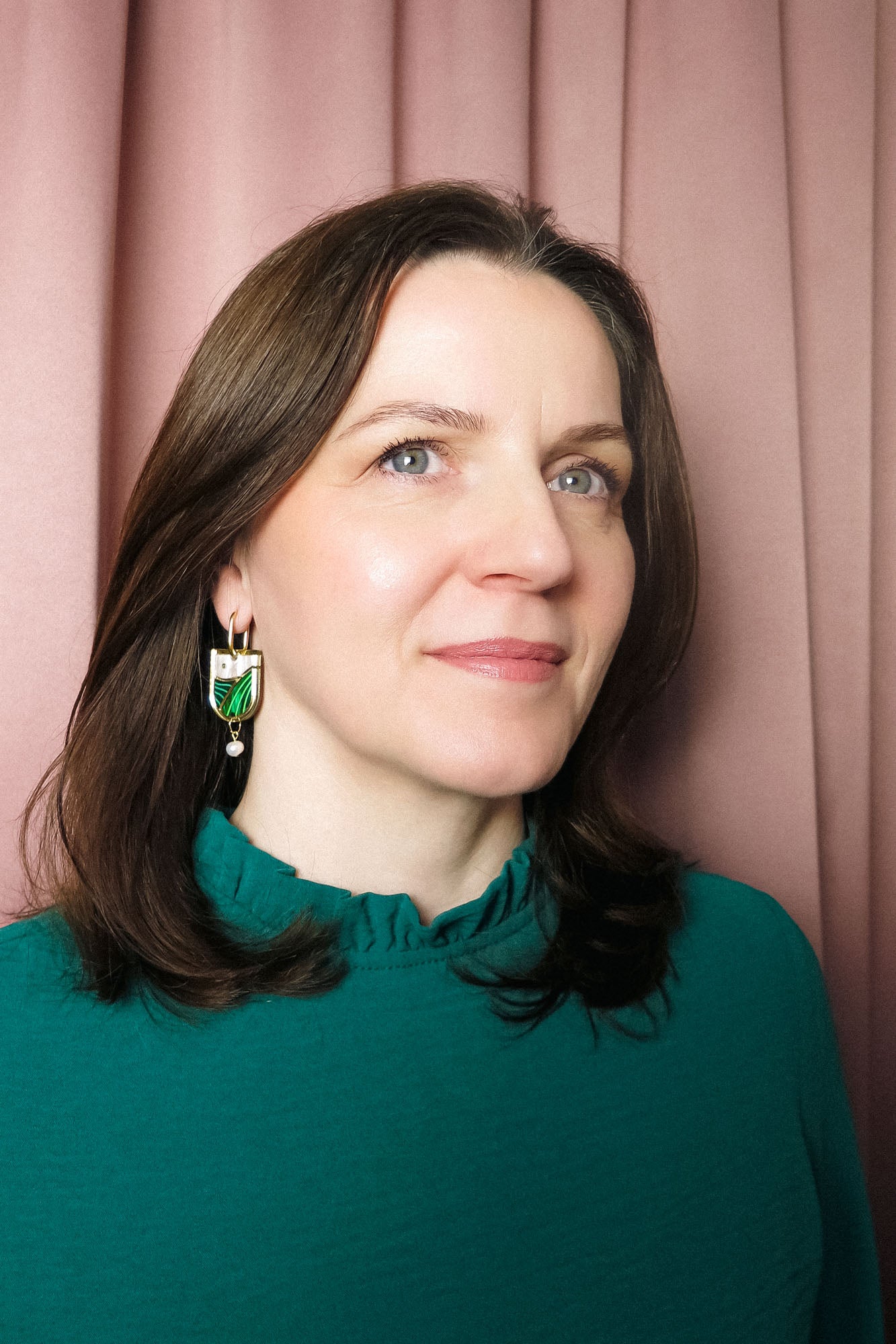 A woman with shoulder-length brown hair, wearing a teal top with a ruffled collar and elegant Vineyard Inspired Art Nouveau Dangle Earrings adorned with pearls & rhinestones, stands in front of pink curtains, looking slightly to the side and smiling softly.