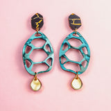 A pair of colorful, sustainable earrings with black and gold geometric tops, teal cut-out oval centers, and small teardrop-shaped gold and white dangles, displayed on a pink background—perfect for fans of handmade accessories.