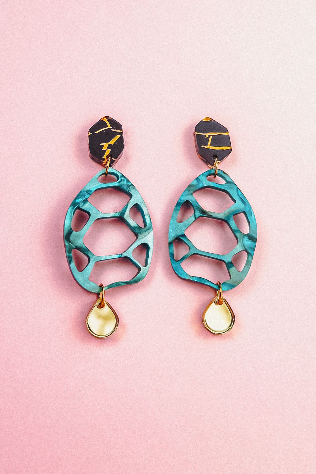 A pair of colorful, sustainable earrings with black and gold geometric tops, teal cut-out oval centers, and small teardrop-shaped gold and white dangles, displayed on a pink background—perfect for fans of handmade accessories.