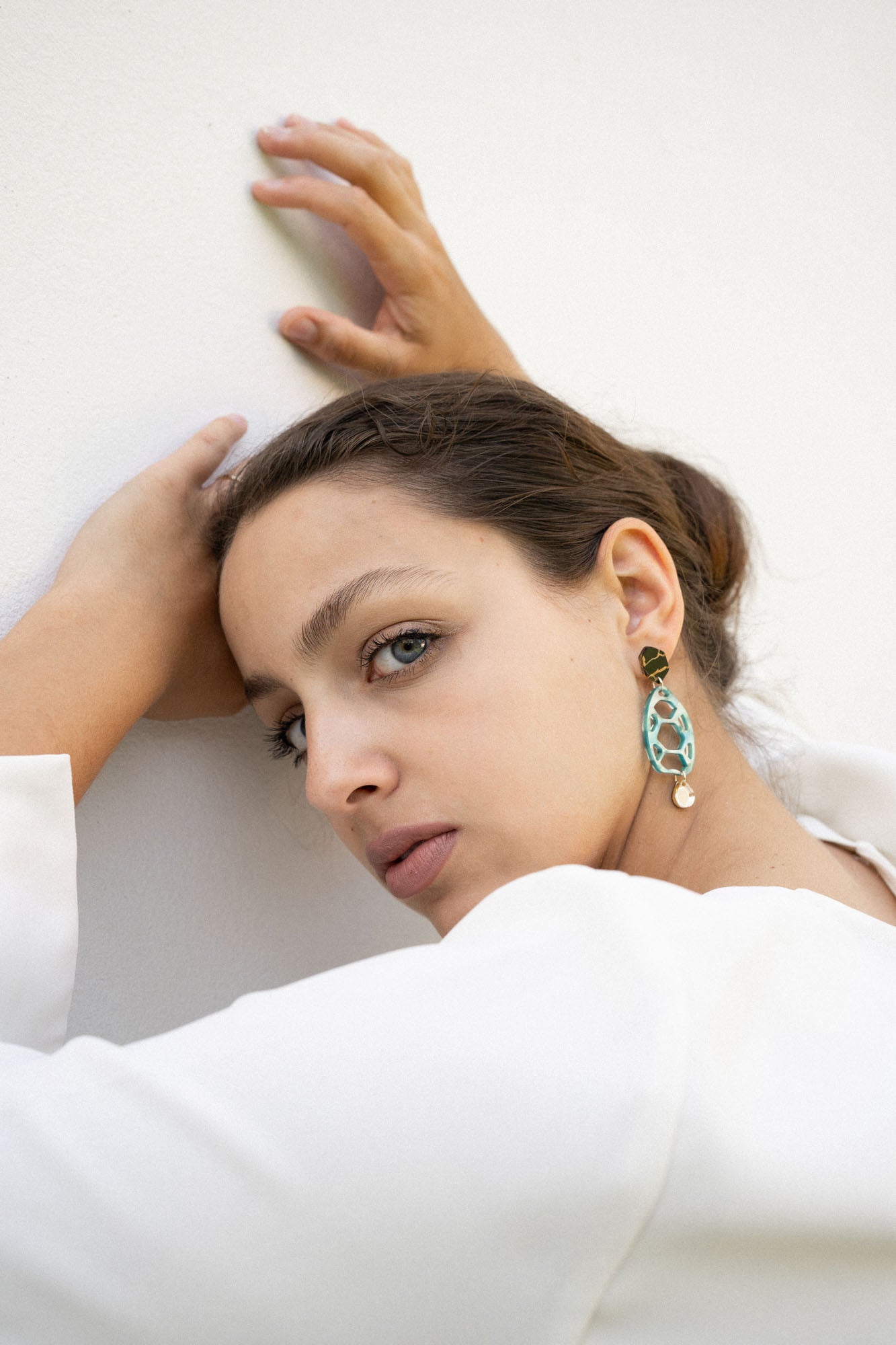 A woman with light eyes and brown hair pulled back leans against a white wall, gazing at the camera. She wears a white top and large, oval sustainable earrings, her hands resting above her head in an effortlessly chic pose.