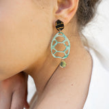 A close-up of a person’s ear wearing a statement, sustainable earring with a black geometric stud, a large mint green oval with cutouts, and a small gold disc at the bottom—showcasing zero-waste beauty. The person has light skin and dark brown hair.