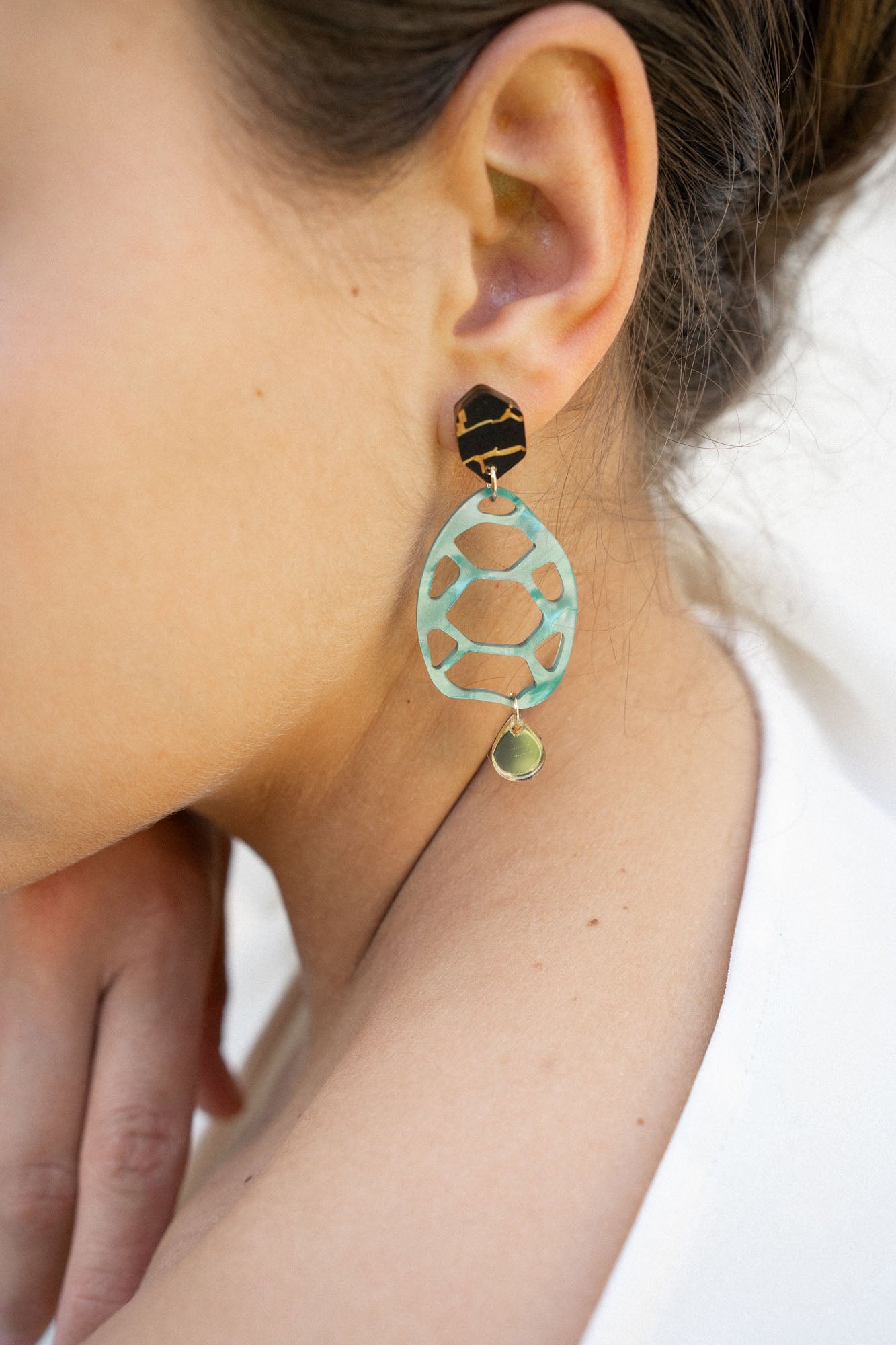 A close-up of a person’s ear wearing a statement, sustainable earring with a black geometric stud, a large mint green oval with cutouts, and a small gold disc at the bottom—showcasing zero-waste beauty. The person has light skin and dark brown hair.