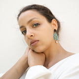 A woman with light eyes and brown hair, wearing a white top, rests her chin on her hand. She wears sustainable turquoise and gold earrings and looks calmly into the camera against a plain white background.