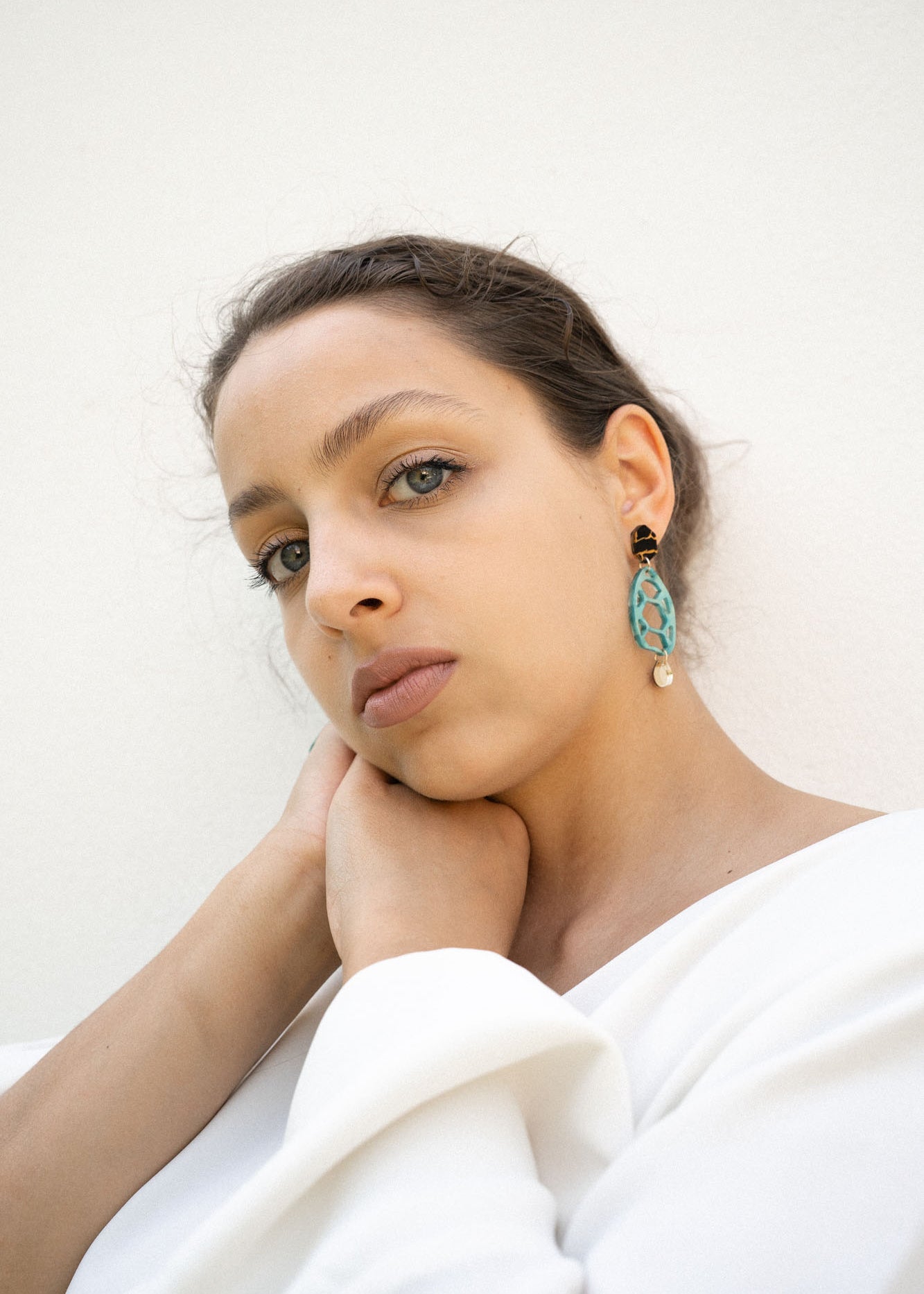 A woman with light eyes and brown hair, wearing a white top, rests her chin on her hand. She wears sustainable turquoise and gold earrings and looks calmly into the camera against a plain white background.