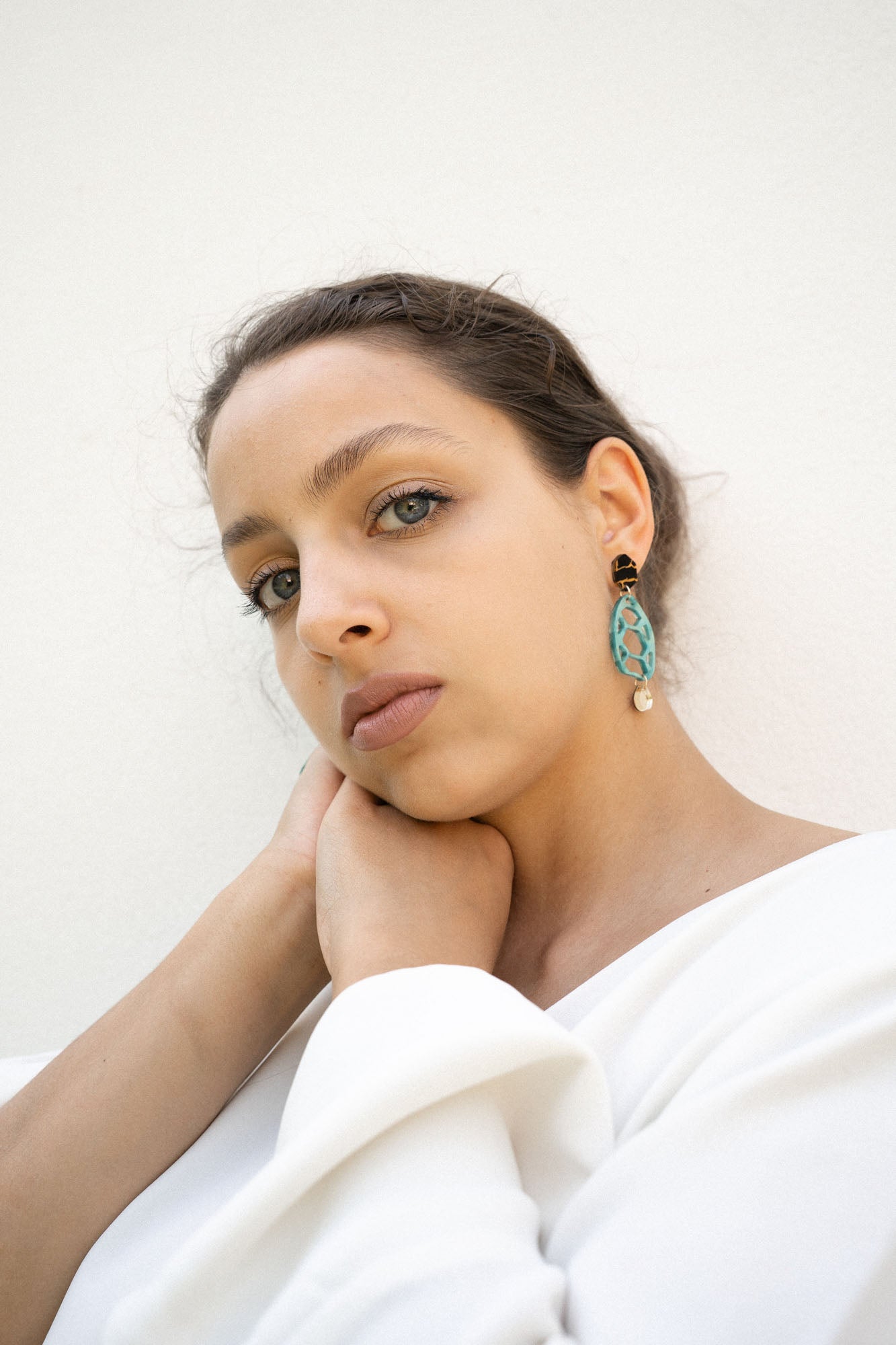 A woman with light eyes and brown hair, wearing a white top, rests her chin on her hand. She wears sustainable turquoise and gold earrings and looks calmly into the camera against a plain white background.
