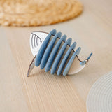A decorative object resembling a stylized fish, made of alternating blue-grey felt and white pieces, sits on a light wooden table with woven placemats in the background, adding home charm and protecting surfaces like unique Whale Coasters.