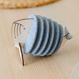 A wooden and felt napkin holder shaped like a fish sits on a light wooden table, adding home charm, with a woven placemat blurred in the background. Pair it with Whale Coasters to protect surfaces and complete your decor.