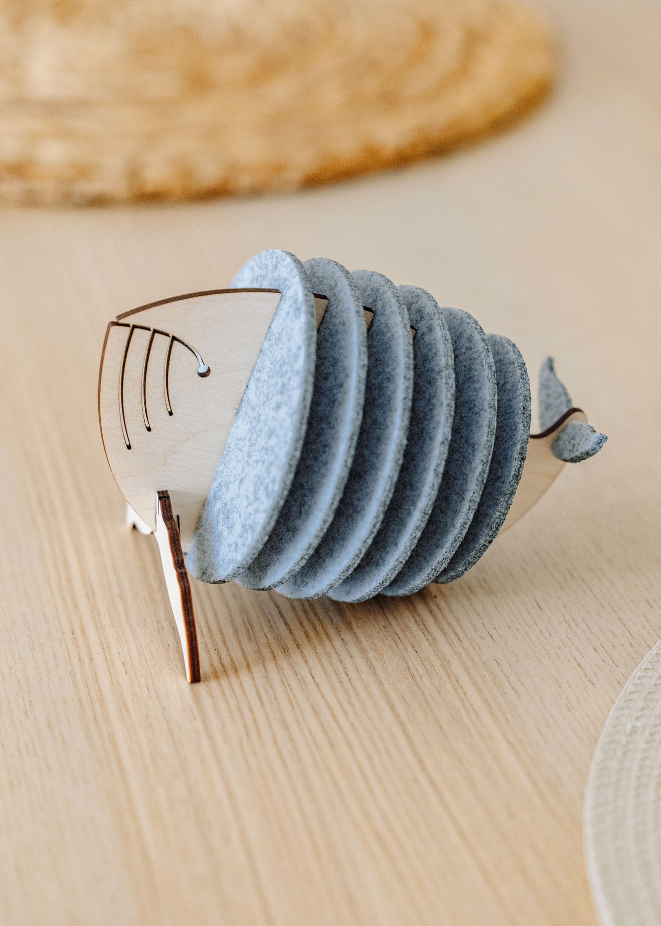 A wooden and felt napkin holder shaped like a fish sits on a light wooden table, adding home charm, with a woven placemat blurred in the background. Pair it with Whale Coasters to protect surfaces and complete your decor.