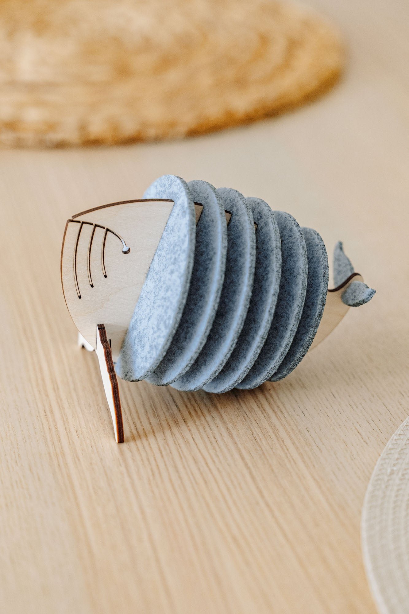 A wooden and felt napkin holder shaped like a fish sits on a light wooden table, adding home charm, with a woven placemat blurred in the background. Pair it with Whale Coasters to protect surfaces and complete your decor.